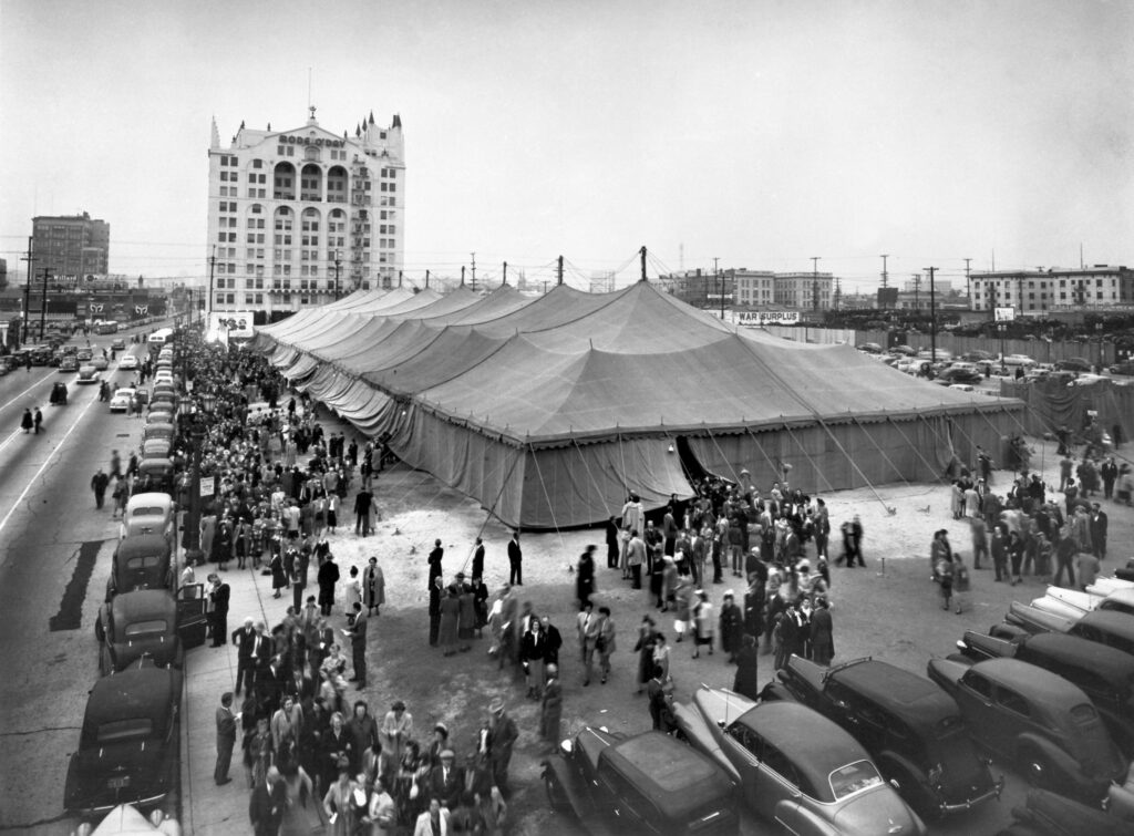 Historic black and white photo of a crowded event with a large tent and a tall building nearby.
