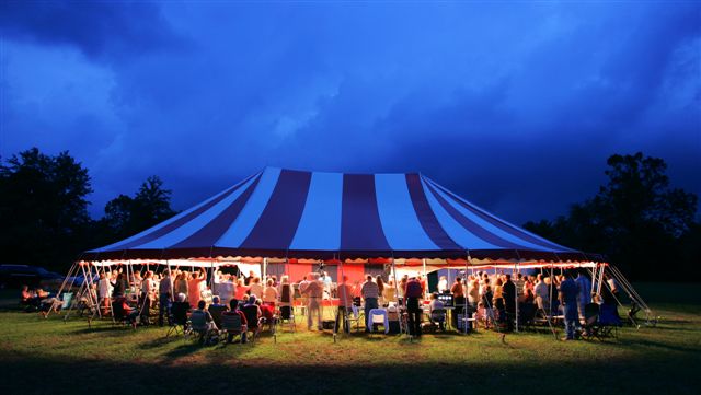 A large striped tent lit warmly at dusk with people gathering around.