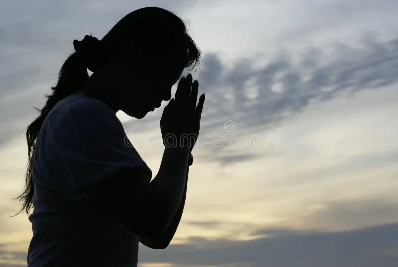 Silhouette of a person praying at sunset with hands clasped.